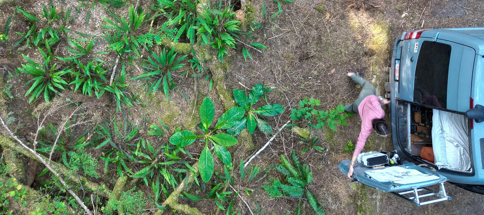 A woman closing the gear garage of a Mercedes 2016 Sprinter Van conversion backed up to coastal rainforest viewed from the top down
