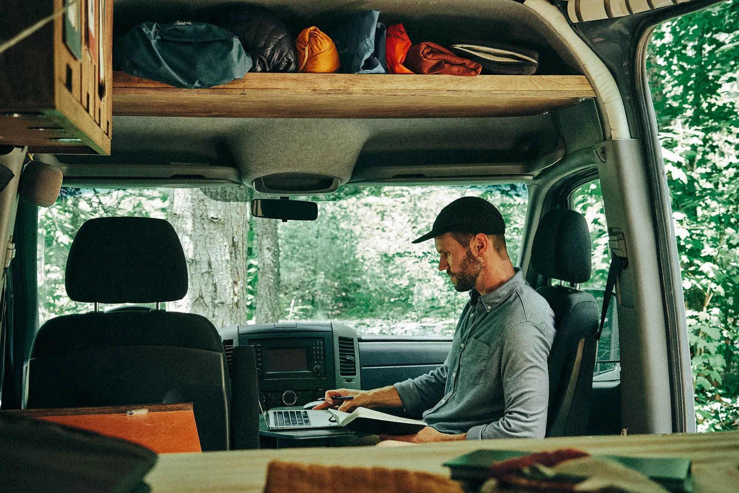 Man working at his computer from the passenger seat of a 2016 mercedes sprinter van below a Vancillary headliner shelf made with the DIY Conversion kit