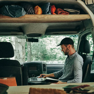 Man working at his desk in the front cabin of a mercedes sprinter conversion van with storage above the front seats visible due to the DIY headliner shelf being installed on this model year and creating highly accessible storage